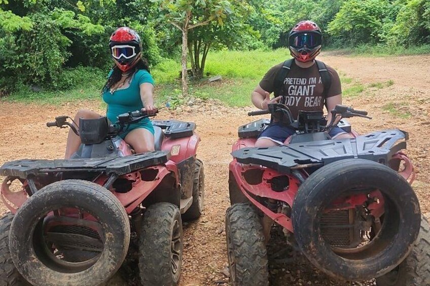 Two people on ATVs in a dirt path with greenery in the background. 3