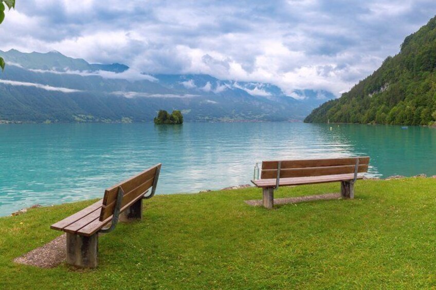 A lake with two wooden benches on the grass, surrounded by mountains and a cloudy sky. 4