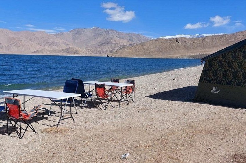 A lakeside campsite with tables and chairs, a tent, and mountains in the background. 1