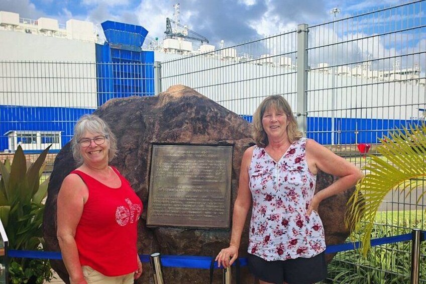 Two people standing in front of a large rock with a plaque, a fence, and industrial buildings in the background. 5
