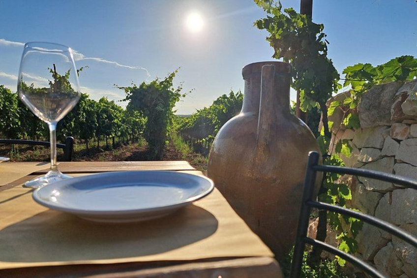 A table with a wine glass and a plate, a large vase, and a vineyard in the background. 5