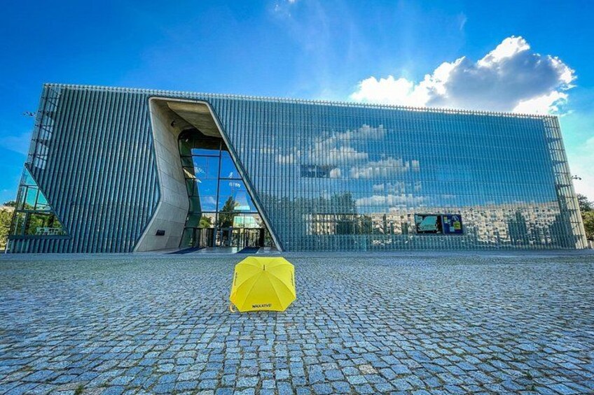 Een modern gebouw met een uniek, hoekig ontwerp, een geplaveid voetpad en een helderblauwe lucht. 1