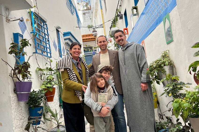 Eine Gruppe von Menschen steht in einem schmalen Gang mit weißen Wänden und blauen Fensterläden, umgeben von Blumentöpfen. 5