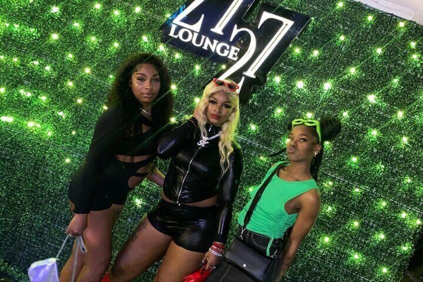 Three people posing in front of a green glittery backdrop with the text "Lounge 21". 1