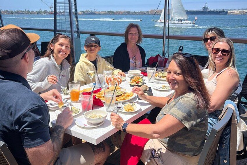 A group of people dining on a boat with a view of the sea and a sailboat in the background. 2