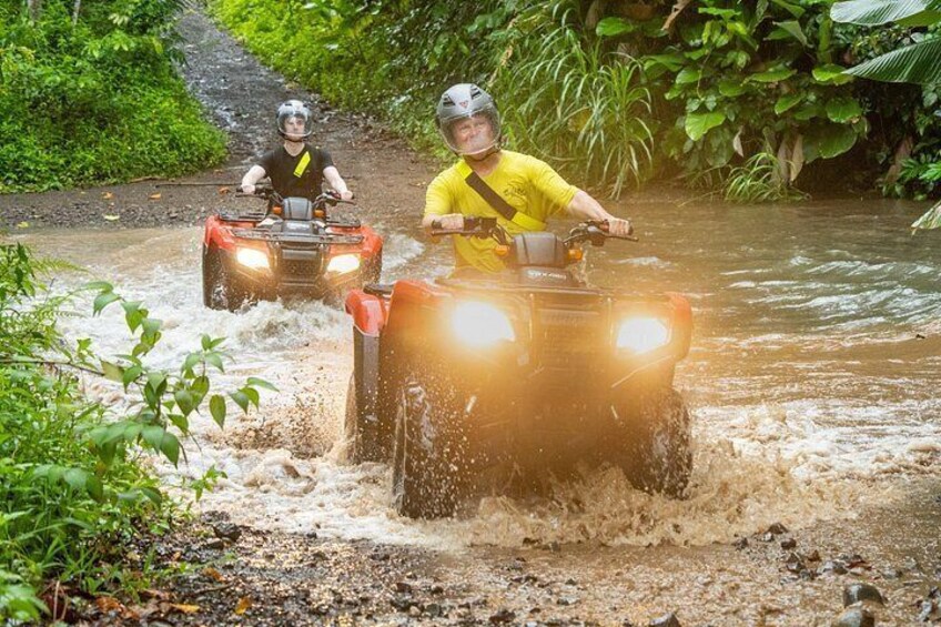 Two people on ATVs navigating a flooded path surrounded by greenery. 2