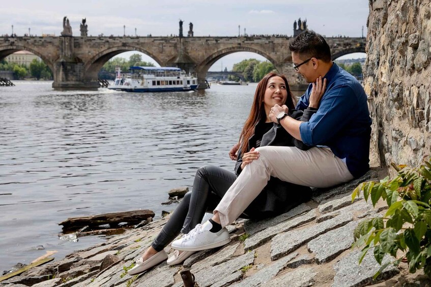 A couple sitting on a stone ledge by a river with a bridge in the background. 1
