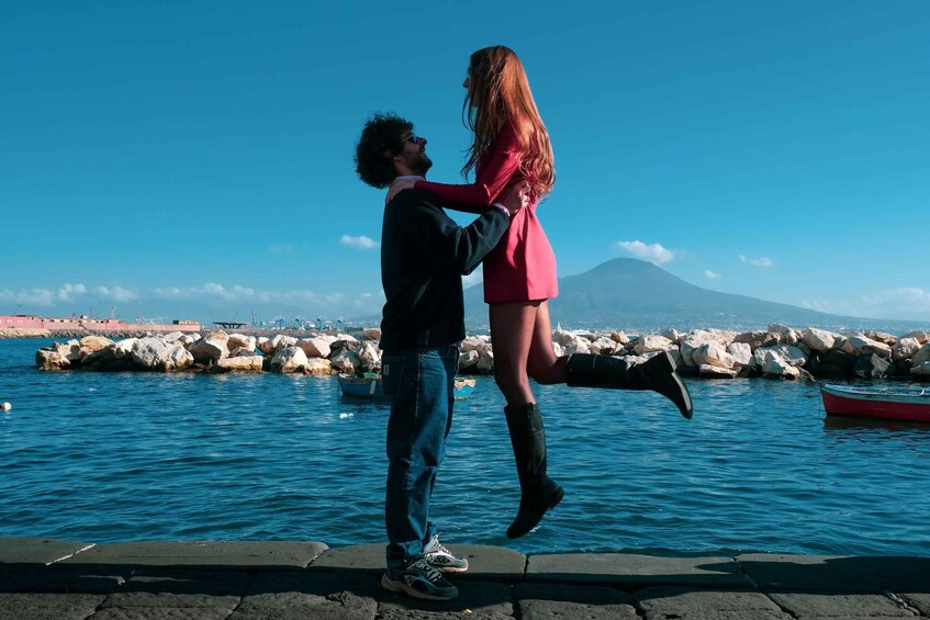 A couple embracing by a waterfront with a rocky breakwater and a boat in the background. 5