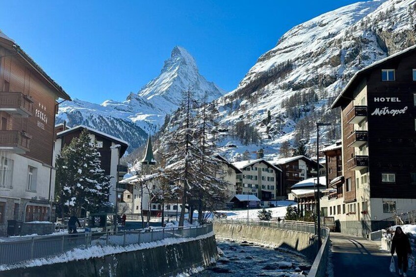 A snow-covered town with a prominent mountain in the background, Hotel Astropal, and a river running through the center. 5