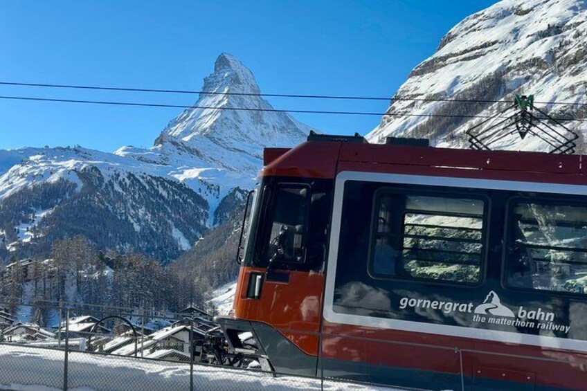 A red train with a sign reading 'gornergratbahn the matterhorn railway' against a backdrop of snow-covered mountains and a clear blue sky. 4