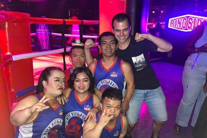 A group of people in a dimly lit indoor setting with a boxing ring and a neon sign. 5