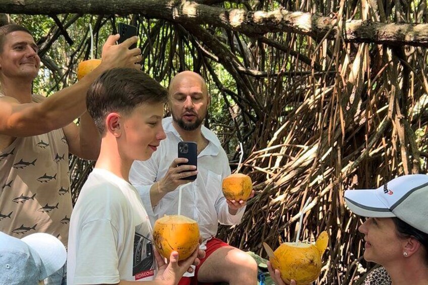 People holding coconuts and taking photos in a natural setting with a tree structure in the background. 2