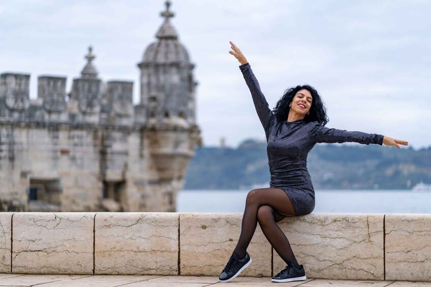 A person sitting on a stone ledge with arms outstretched, overlooking a historic building and a body of water. 4