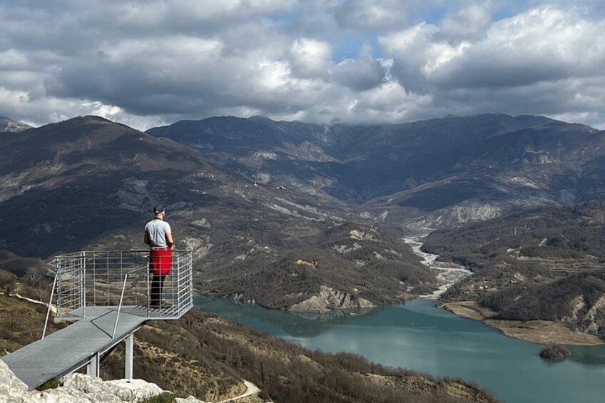 A person standing on a metal platform overlooking a river valley with mountains in the background. 2