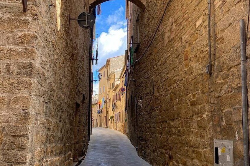 A narrow stone alleyway flanked by buildings with visible windows and doors, leading to a street with a clear sky above. 3