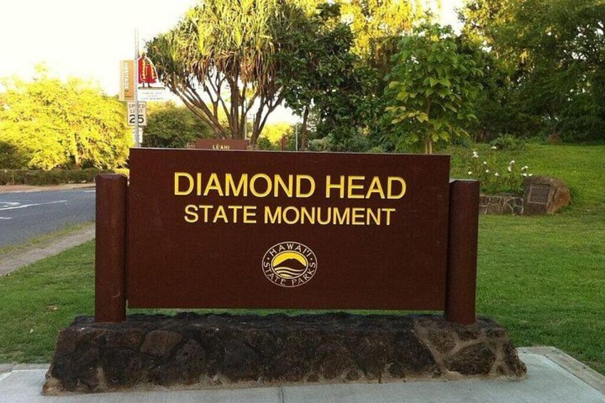 Een bord voor het Diamond Head State Monument met een logo, tegen een achtergrond van bomen en een weg. 5