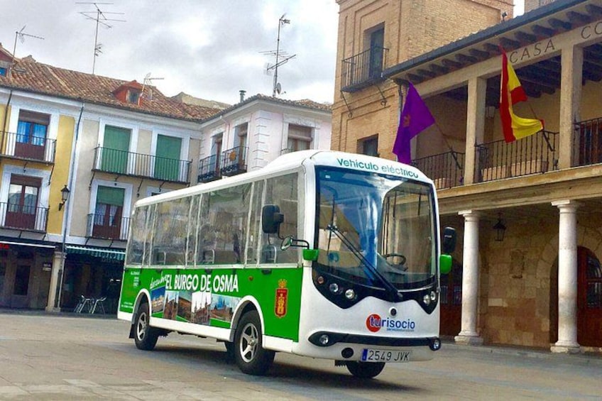 A white and green bus with 'El Burgo de Osma' and 'Turisocio' on its side, parked in front of a building with balconies and flags. 3