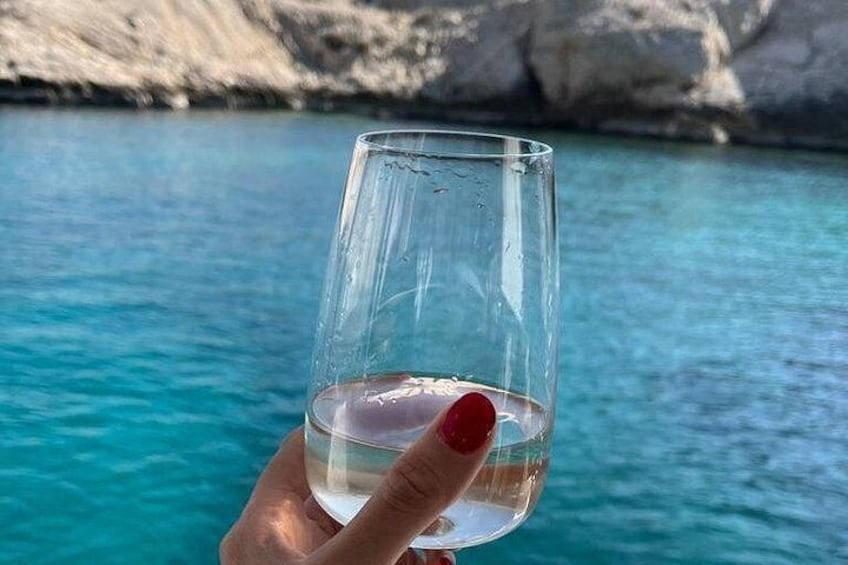 A hand holding a clear glass with a liquid, against a backdrop of turquoise water and rocky cliffs. 5