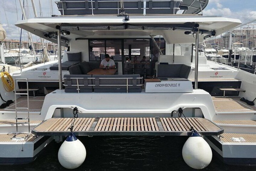 A yacht named 'GARAMBIOLLE II' docked at a marina with other boats, a wooden deck, and a seating area. 4