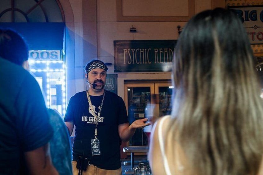 A person in a black t-shirt with a gold chain standing in a room with a sign that reads 'PSYCHIC READINGS'. 1