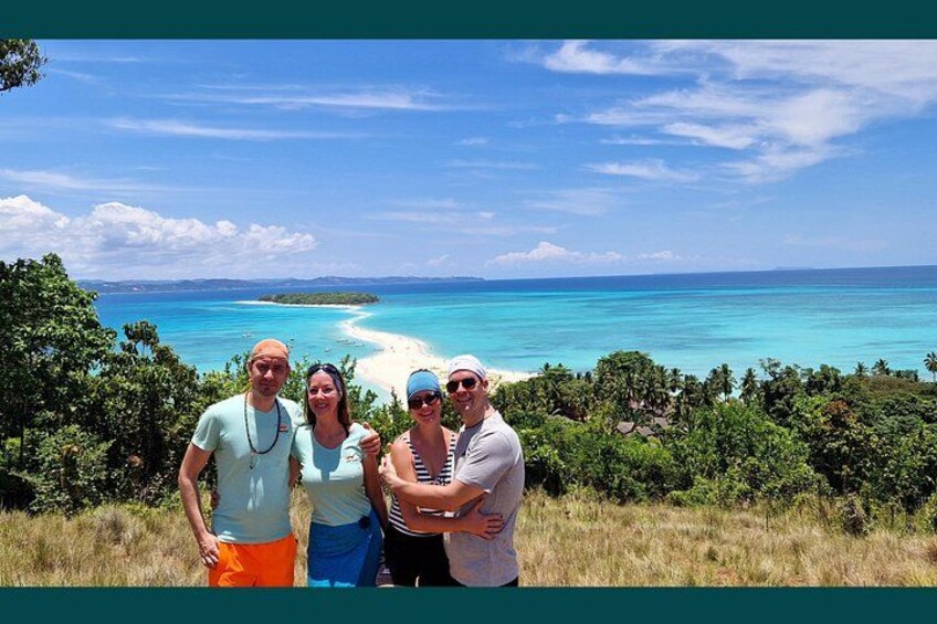 A group of people standing on a grassy hill overlooking a beach with clear turquoise waters and a distant shoreline. 3