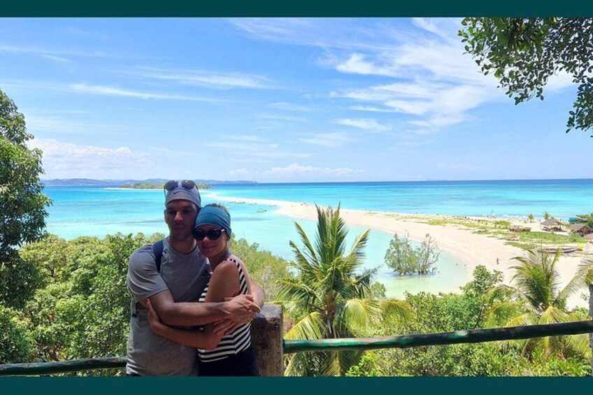 A couple embracing on a balcony overlooking a turquoise beach with palm trees and a clear sky. 5