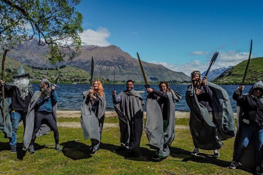 A group of people in medieval costumes holding swords by a lake with mountains in the background. 4
