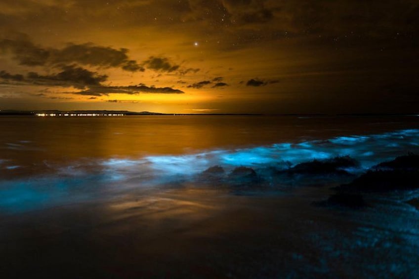 Escena costera nocturna con puesta de sol, olas y un horizonte urbano lejano. 2
