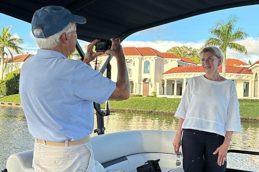 Un hombre tomando una foto con una cámara, una mujer a su lado, y un barco al fondo con una casa y palmeras. 1