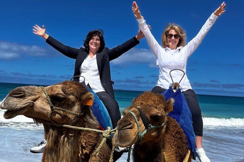Two people riding camels on a beach with the ocean in the background. 1