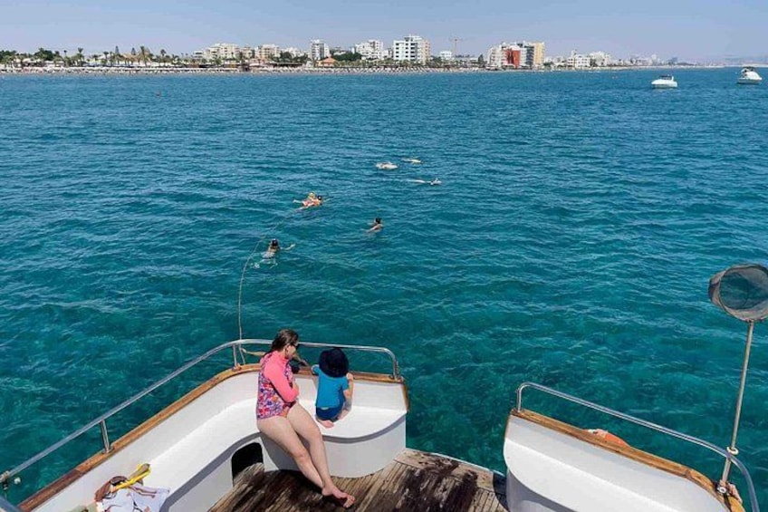 A boat with two people on the deck, a clear blue sea, and a cityscape in the background. 2
