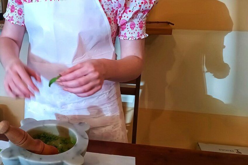 A person wearing a white apron and pink floral shirt, holding a green leaf, standing next to a mortar and pestle with a bowl of green mixture. 2