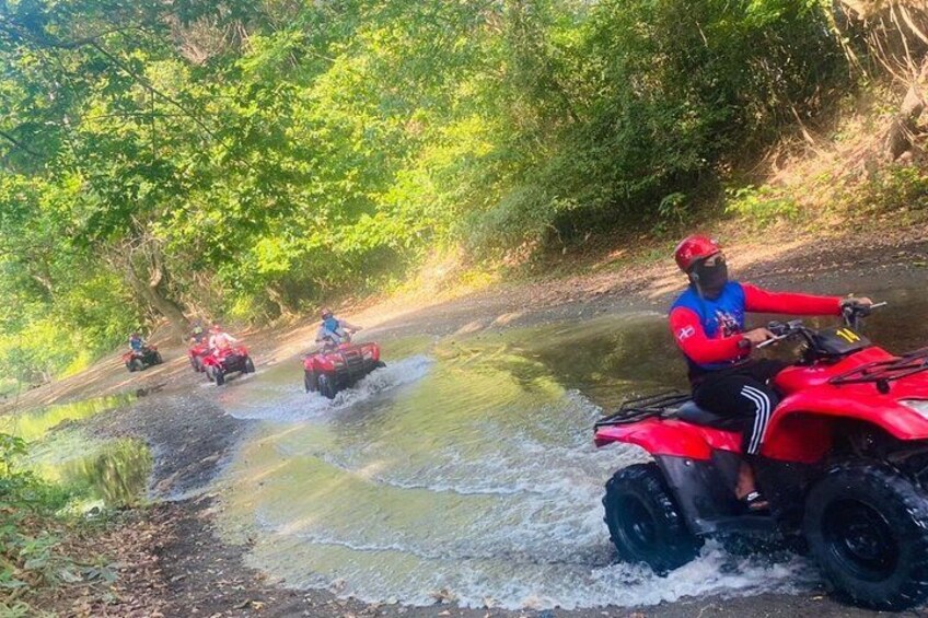 Une personne conduit un quad rouge sur un étroit passage d’eau, entouré d’une végétation luxuriante. 2