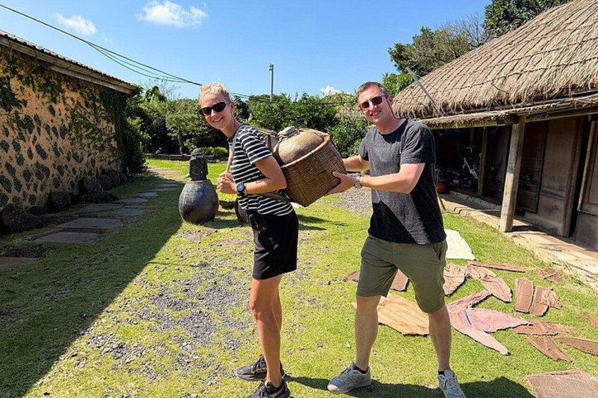 Two people carrying a large woven basket, a stone pot, and a thatched-roof structure in a garden. 2