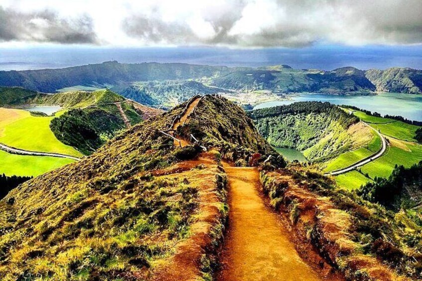 A mountain trail leading to a valley with a lake and distant hills. 1