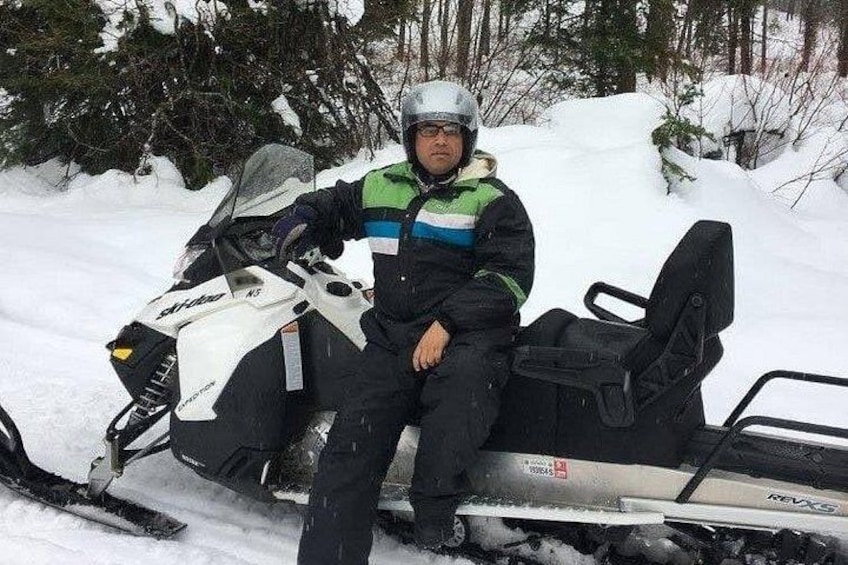 Une personne assise sur une motoneige, portant un casque, entourée de neige et d’arbres. 2