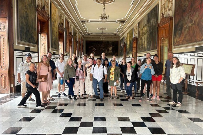Een groep mensen staat in een statige hal met een geruite vloer, grote schilderijen aan de muur en een kroonluchter. 2