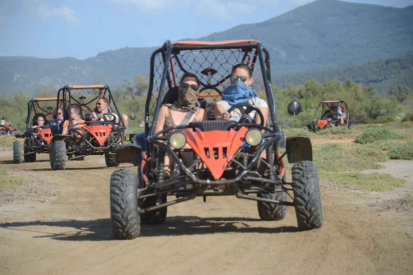 A group of people in off-road vehicles on a dirt road with mountains in the background. 1