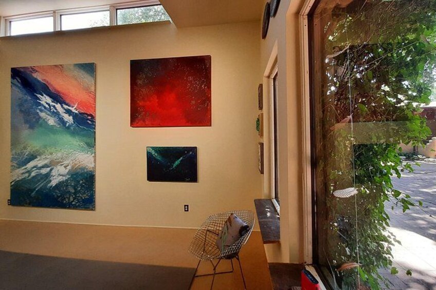 A room with three large paintings on the wall, a chair, and a window with greenery outside. 5