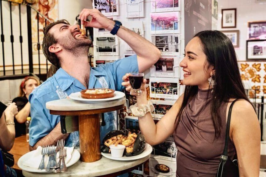 A person eating a dessert with a fork and knife, and another person holding a drink. 1