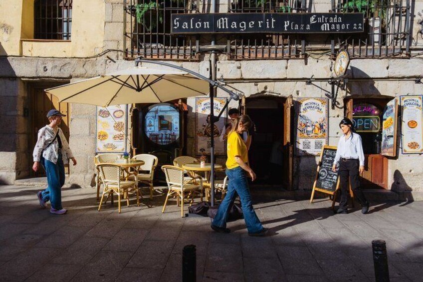 A street with outdoor seating, a sign for 'Salon Magerit Plaza Mayor 22,' and people walking by. 5