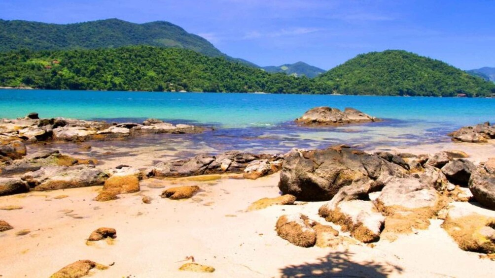 Une plage à l’eau turquoise et limpide, avec un rivage rocheux et des collines verdoyantes en arrière-plan. 2