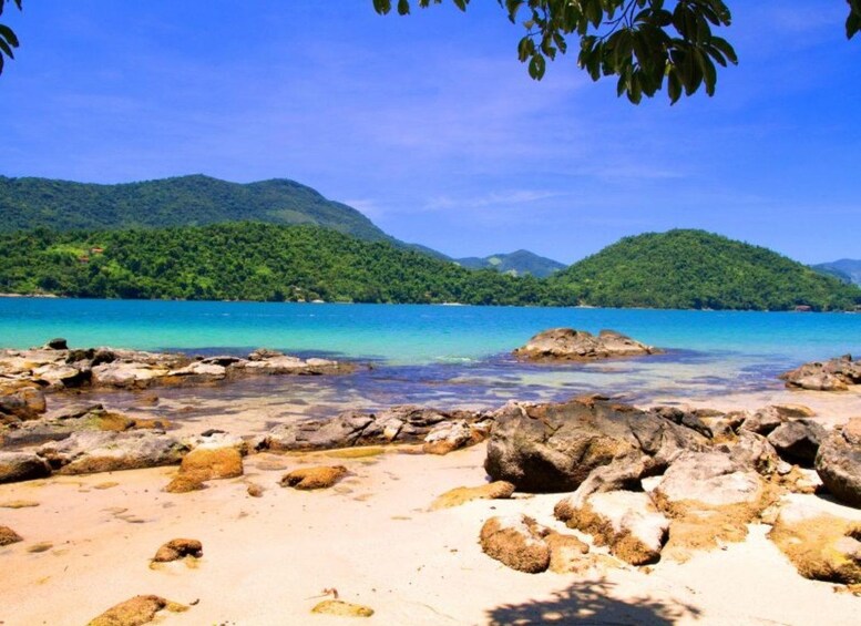 A beach with clear turquoise water, rocky shore, and lush green hills in the background. 2