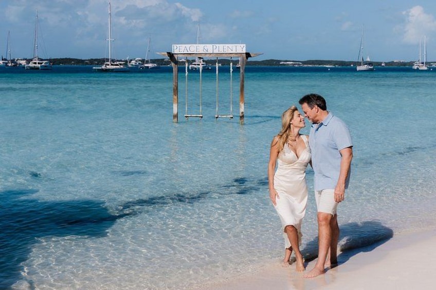 A couple standing on a beach with clear water, a sign reading 'PEACE & PONY,' and sailboats in the background. 1