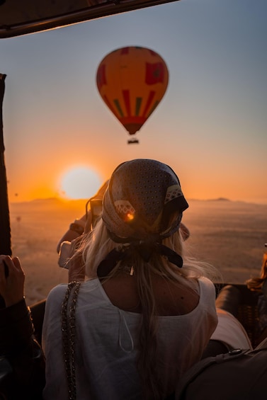 A person with a helmet and a white top, looking at a hot air balloon in the distance during sunset. 2
