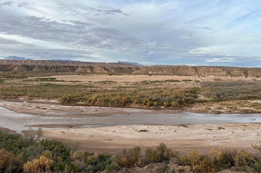 A dry riverbed with sparse vegetation and distant mountains under a cloudy sky. 3