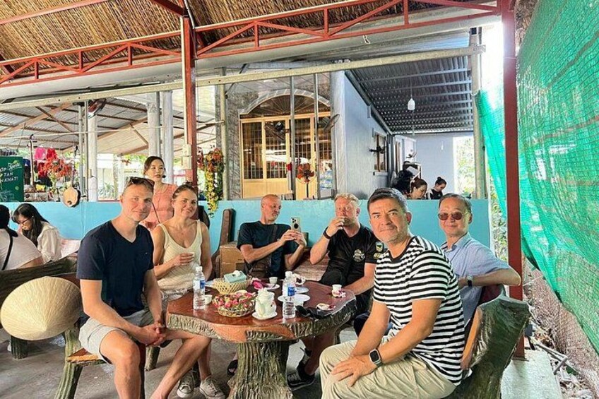 A group of people sitting at an outdoor table with drinks and food, under a covered area with a thatched roof. 2