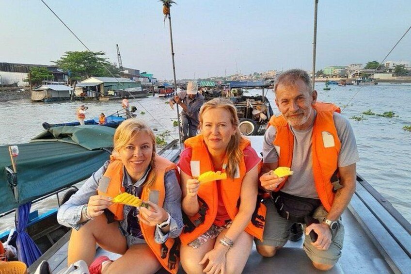 Three people on a boat wearing orange life jackets, holding yellow objects, with a waterway and other boats in the background. 5