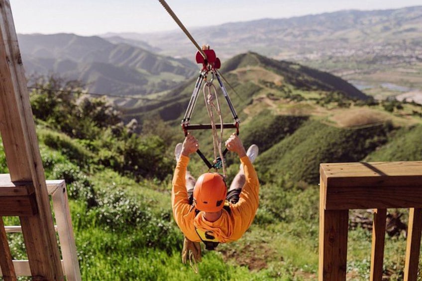 A person in an orange shirt hanging from a rope swing, suspended above a scenic landscape with hills and greenery. 3