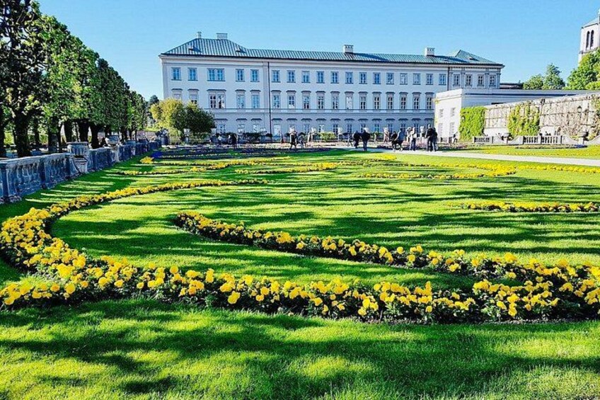 Ein großes Gebäude mit einer symmetrischen Fassade, einem gepflegten Rasen mit gelben Blumen und einer Steinmauer im Hintergrund. 4